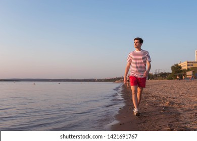 A handsome, young guy, a man in a white T-shirt with stripes and shorts is walking along the embankment of the river, the sea. Walking at the resort. Rest by the beach.