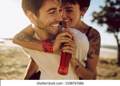 Handsome Young Guy Piggybacking His Girlfriend Drinking Soft Drink Outdoors. Happy Caucasian Couple Enjoying Themselves Outdoors.