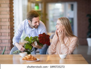 Handsome Young Guy Presenting Bouquet Of Flowers To His Excited Girlfriend At Cafe, Making Her Special Surprise. Loving Couple Celebrating Valentine's Day, Birthday Or Anniversary