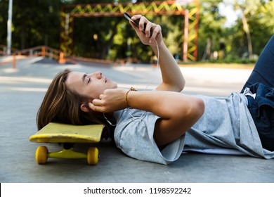 Handsome young guy spending time at the skate park, listening to music with headphones, laying on skateboard, holding mobile phone - Powered by Shutterstock