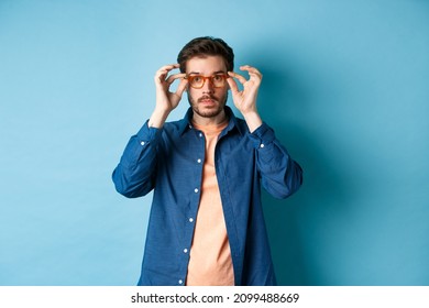 Handsome Young Guy Trying New Glasses In Store, Looking At Camera, Standing On Blue Background