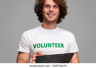 Handsome Young Guy In Volunteer T-shirt Smiling And Looking Away While Standing And Holding Clipboard With Petition On Gray Background