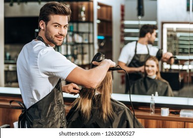 handsome young hairstylist smiling at camera while drying hair to beautiful young woman in beauty salon 
