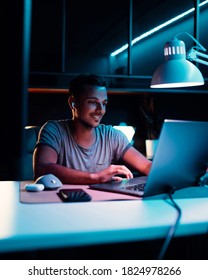 Handsome Young Happy IT Specialist Uses Computer, Browses In Internet, Plays Online Games, Does Hacking. Cool Neon Hue Lights In His Office Room.