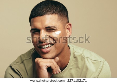Handsome young having fun while taking care of his skin, he smiles and winks at the camera with beauty cream on his face. Happy young man using a nourishing product in his skincare routine.