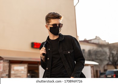 Handsome Young Hipster Man With Hairstyle In Trendy Sunglasses In Fashionable Black Denim Jacket In Stylish Medical Black Mask Walks On Street On Sunny Day. Attractive Guy Model. Fashion 2020. Spring.