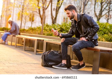 A handsome young hipster man sitting on a bench in the sunset and using his smartphone