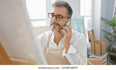 Handsome young hispanic artist with grey hair, engrossed in a vibrant phone conversation while confidently drawing at his bustling art studio. - Powered by Shutterstock