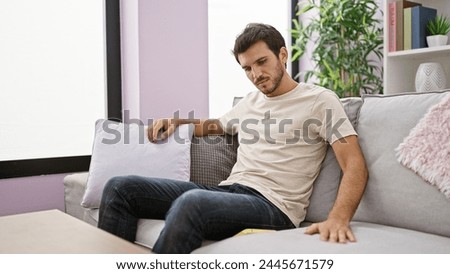 A handsome young hispanic man with a beard relaxes thoughtfully in a modern living room, personifying casual comfort.
