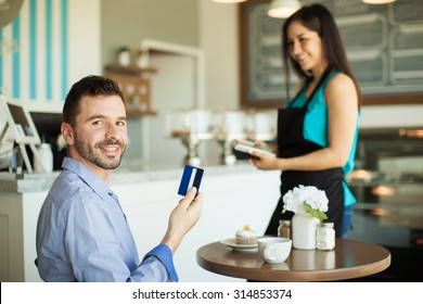 Handsome Young Hispanic Man Holding A Credit Card After Paying For His Coffee And Cupcake At A Cafe And Smiling