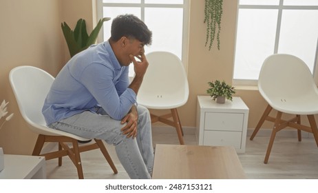 Handsome young hispanic man yawning in a bright, modern waiting room, sitting on a white chair with a thoughtful expression, surrounded by minimalist decor and indoor plants. - Powered by Shutterstock