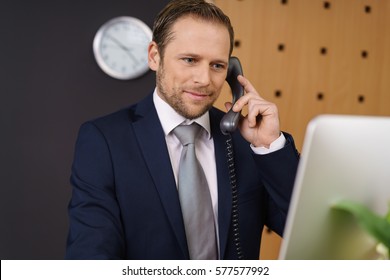 Handsome Young Hotel Manager Checking Information On His Desktop Computer At The Front Reception Desk As He Listens To A Call From A Customer