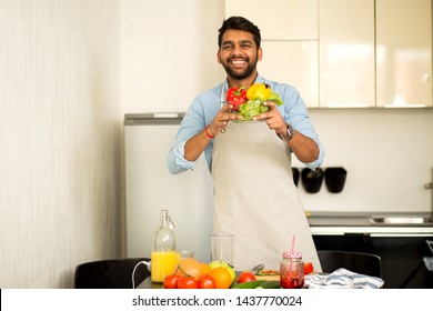 Handsome Young Indian Man In Blue Shirt And Apron Looking At Camera And Smiling While Standing In Kitchen At Home, Preparing Vegetable Salad, Taking Care Of His Health. Healthy Food, Cooking Concept.