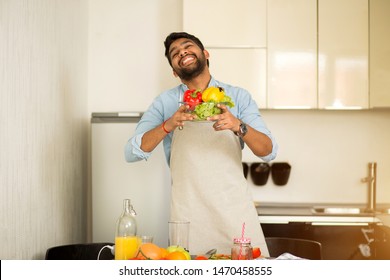 Handsome Young Indian Man In Blue Shirt And Apron Looking At Camera And Smiling While Standing In Kitchen At Home, Preparing Vegetable Salad, Taking Care Of His Health. Healthy Food, Cooking Concept.