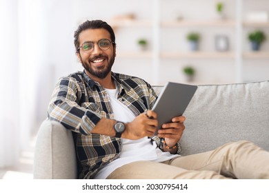 Handsome Young Indian Man Chilling At Home With Digital Tablet, Sitting On Couch, Closeup, Copy Space. Happy Guy Freelancer Shopping Online, Using Modern Pad And Smiling, Entertainment Concept