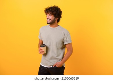 Handsome young indian man with curly hairstyle in casual t-shirt enjoying coffee to-go standing isolated on yellow background, smiling guy with a paper cup of hot drink
