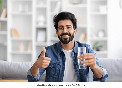 Handsome Young Indian Man Holding Glass Of Water And Showing Thumb Up Gesture At Camera While Sitting On Couch At Home, Smiling Eastern Guy Recommending Healthy Refreshing Drink, Copy Space - Powered by Shutterstock