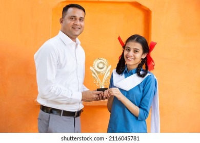 Handsome young Indian man school teacher give victory trophy to girl student against orange background, braided female receive winning prize, skill india. child dreams and support. Education concept.