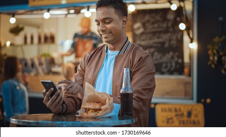 Handsome Young Indian Man is Using a Smartphone while Sitting at a Table in a Outdoors Street Food Cafe. He's Browsing the Internet or Social Media, Posting a Status Update. Man is Happy and Smiling. - Powered by Shutterstock