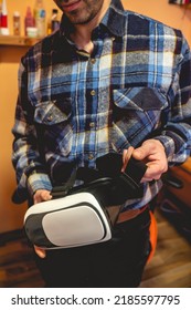 Handsome Young Latino Man With Plaid Shirt Holding A Virtual Reality Helmet (VR Glasses) In Orange Bedroom On Home