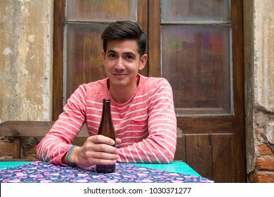 A Handsome Young Latino Man In A Stylish Striped Sweater And Modern Hairstyle, Holds A Brown Beer Bottle At A Table In A Rustic Hipster Dive Bar Or Cantina, Looking To Camera. With Room For Copy Text.