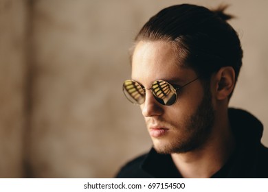 Handsome Young Looking Guy Wearing Black Jacket And Sunglass. There Is Reflection Of The Windows In The Sunglasses. Slight Beard With Little Moustache. Pony Tail On Backside Is Visible.