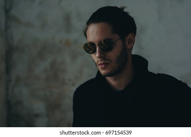 Handsome Young Looking Guy Wearing Black Jacket And Sunglass. There Is Reflection Of The Windows In The Sunglasses. Slight Beard With Little Moustache. Pony Tail On Backside Is Visible.