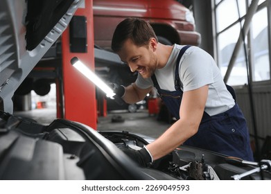 Handsome young male auto mechanic in special uniform clothes holding a flashlight, looking for breakdown and repairing under the hood in the car engine in a car workshop. - Powered by Shutterstock