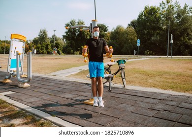 Handsome Young Male Caucasian Athlete With Face Mask Trains On Arm And Leg Equipment In An Outdoor Gym. Muscular Athlete Outdoors. COVID - 19 Coronavirus Protection