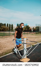 Handsome Young Male Caucasian Athlete With Face Mask Trains On Arm And Leg Equipment In An Outdoor Gym. Muscular Athlete Outdoors. COVID - 19 Coronavirus Protection