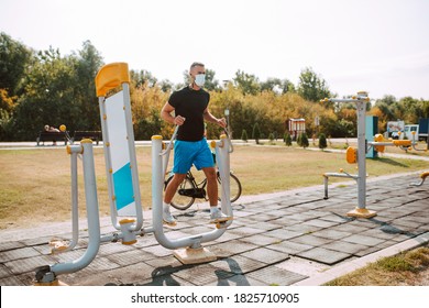 Handsome Young Male Caucasian Athlete With Face Mask Trains On Arm And Leg Equipment In An Outdoor Gym. Muscular Athlete Outdoors. COVID - 19 Coronavirus Protection