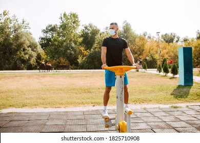 Handsome Young Male Caucasian Athlete With Face Mask Trains On Arm And Leg Equipment In An Outdoor Gym. Muscular Athlete Outdoors. COVID - 19 Coronavirus Protection