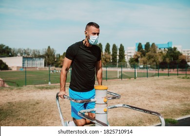 Handsome Young Male Caucasian Athlete With Face Mask Trains On Arm And Leg Equipment In An Outdoor Gym. Muscular Athlete Outdoors. COVID - 19 Coronavirus Protection