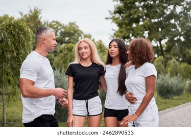 Handsome young male coach talking with diverse women in sportswear at park. Multiethnic fit female friends listening to coach man during training session in nature. - Powered by Shutterstock