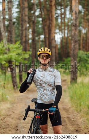 Handsome young male cyclist stands in the woods with a bottle of water in his hand and looks at the camera.