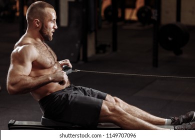 Handsome Young Male Pulling The Indoor Rower. Side View Photo. Development Of Muscles