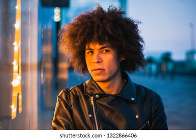 Handsome young man with afro hair illuminated by yellow light of a showcase looking seriously with urban background in blue tones - Powered by Shutterstock