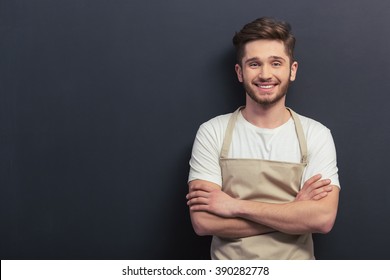 Handsome Young Man In Apron Is Looking At Camera And Smiling, Standing With Crossed Arms Against Blackboard