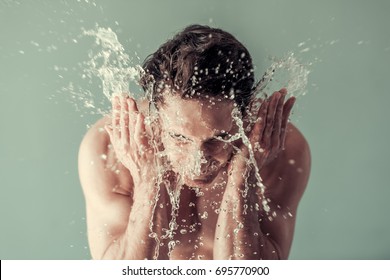 Handsome young man with bare torso is washing his face, splashes of water around, gray background