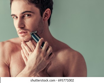 Handsome young man with bare torso is using an electric shaver and looking at camera, on gray background