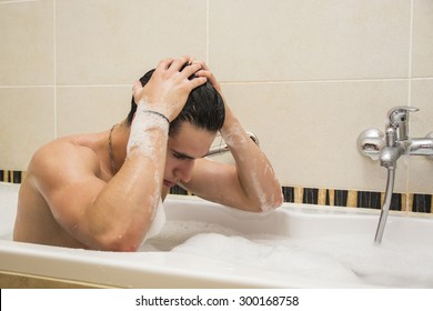 Handsome young man in bathtub at home having bath, washing body and hair with bathfoam and shampoo