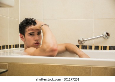 Handsome young man in bathtub at home having bath, leaning on tub edge, looking at camera