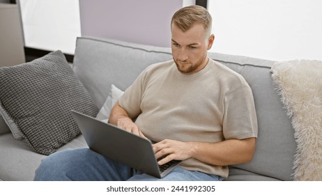A handsome young man with a beard comfortably using a laptop on a sofa in a modern living room setting.