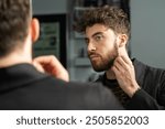 Handsome young man with beard looking in mirror, touching his face. Concept of male grooming, self-care routine, and modern masculinity. Stylish haircut and facial hair maintenance.