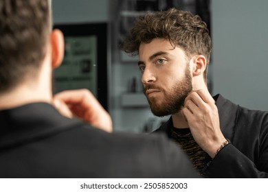 Handsome young man with beard looking in mirror, touching his face. Concept of male grooming, self-care routine, and modern masculinity. Stylish haircut and facial hair maintenance.