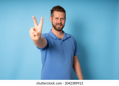 Handsome young man with beard wearing blue polo t-shirt smiling looking to the camera showing fingers doing victory sign.
