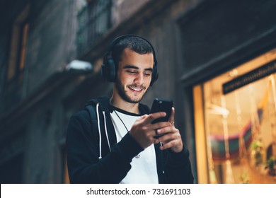 Handsome young man in black hoodie wears big wireless music headphones and changes audio tracks on smartphone, he smiles when looks at phone screen
