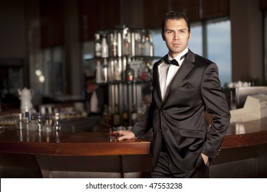 Handsome Young Man In A Black Tuxedo At A Round Bar Holding Whiskey In His Hand; Shallow Depth Of Field, Focus On Face.