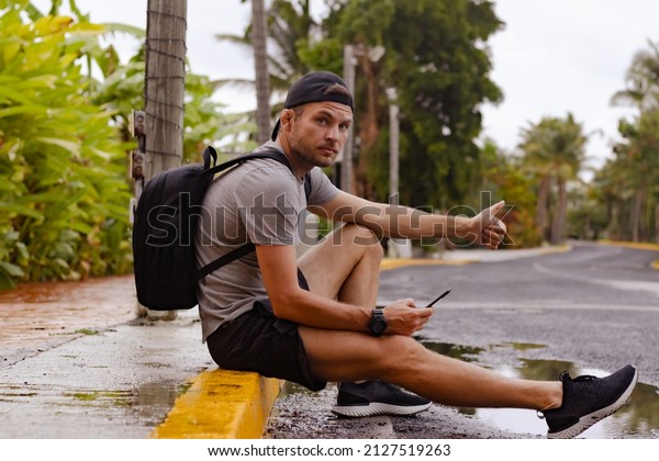 Handsome\
young man in cap with backpack sits on the roadside. Hitchhiker\
catches a car sitting on the road in\
tropic.