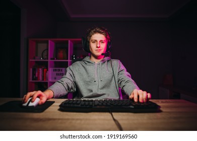 Handsome young man in casual clothes and headset sitting at a table in a room with purple lights and playing video games on the computer with a smile on his face looking at the screen.